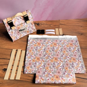 Peut inclure: Un sac à imprimé floral avec une feuille de tissu assortie et des sangles beiges. Le sac a des boucles noires et une poignée beige. La feuille et le sac présentent un motif de fleurs orange, roses et violettes sur fond blanc. Le sac porte le mot "Miam!"