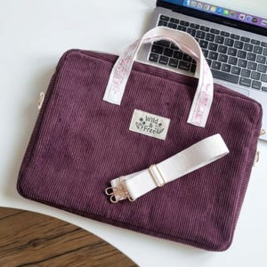 Peut inclure: Une sacoche pour ordinateur portable en velours côtelé violet foncé avec deux poignées claires à motif floral et une bandoulière amovible. Un écusson blanc rectangulaire sur le devant affiche le texte Wild & Free avec une illustration florale.