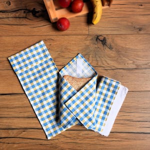 Pode incluir: Um embrulho de sanduíche reutilizável xadrez azul, amarelo e branco com uma fatia de pão dentro. O embrulho está aberto, revelando o pão, e está ao lado de um guardanapo a condizer. O fundo é uma superfície de madeira.