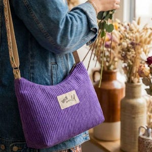 May include: A purple corduroy crossbody bag with a rectangular fabric label featuring the text "hello world" in black script. The bag has a wide, adjustable shoulder strap with a beige and brown floral pattern and a zipper closure along the top.