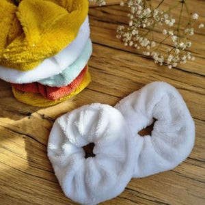 Op de afbeelding: Twee witte, pluche haar elastiekjes op een houten oppervlak. De elastiekjes zijn gemaakt van een zacht, pluche materiaal en zijn perfect om haar op zijn plaats te houden.