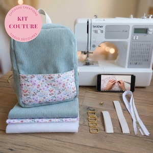 Peut inclure: Un kit de sac à dos en velours côtelé vert sauge avec une poche avant à motif floral, une machine à coudre blanche, un smartphone affichant un tutoriel, quatre boucles rectangulaires dorées, deux sangles blanches et une fermeture éclair blanche.