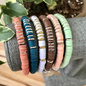 Puede incluir: Un conjunto de seis pulseras coloridas hechas de cuentas redondas apiladas. Las pulseras son en tonos melocotón, azul verdoso, blanco, marrón, rosa y verde claro. Cada pulsera tiene un dije plateado.