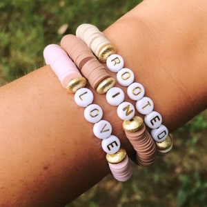 May include: Three beaded bracelets with the words "LOVE", "IN", and "ROOTED" spelled out in gold letters on white beads. The bracelets are layered on a person's wrist and are made of pink, brown, and white beads.