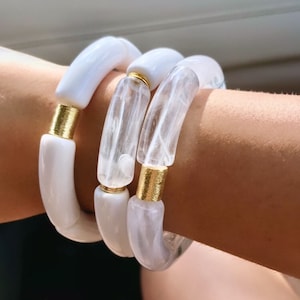 Może przedstawiać: Trzy białe bransoletki typu bangle ze złotymi akcentami. Bransoletki są ułożone na nadgarstku osoby.