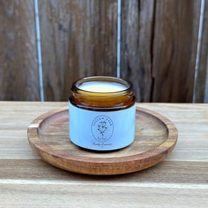 Op de afbeelding: Een klein, amberkleurig glazen potje met Lovely Lavender kaarsenbalsem staat op een ronde, houten schaal. Het potje heeft een lichtblauw etiket met de tekst "Tallow Balm by Lips" en een bloemenillustratie. De achtergrond is een houten oppervlak.