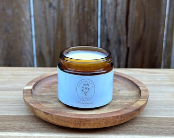 May include: A small, amber-colored glass jar of Lovely Lavender candle balm sits on a round, wooden tray. The jar has a light blue label with the text "Tallow Balm by Lips" and a floral illustration. The background is a wooden surface.