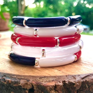 Puede incluir: Pila de cuatro pulseras en colores rojo, blanco y azul. Cada pulsera está hecha de cuentas redondas y lisas con detalles plateados.
