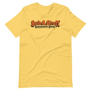 Pode incluir: Camiseta amarela de manga curta com um logotipo de estilo retrô que diz "Sub 8 Stuff Sandwich Shop".