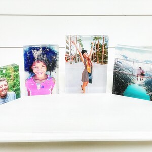 May include: Four clear acrylic photo blocks displaying colorful images. The images include a smiling woman, a young girl in a pink shirt, a graduate in a cap and gown, and a scenic mountain view with a turquoise lake.