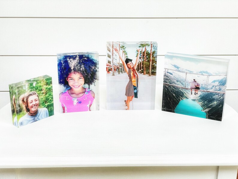 May include: Four clear acrylic photo blocks displaying colorful images. The images include a smiling woman, a young girl in a pink shirt, a graduate in a cap and gown, and a scenic mountain view with a turquoise lake.