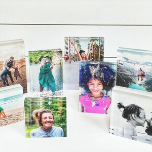 May include: A collection of clear acrylic photo blocks displaying various images. The photos include people, couples, and families in different settings, such as a beach, a graduation, and a mountain landscape. The blocks are arranged on a white surface.
