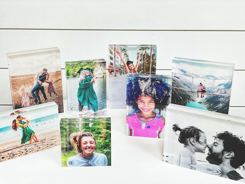 May include: A collection of clear acrylic photo blocks displaying various images. The photos include people, couples, and families in different settings, such as a beach, a graduation, and a mountain landscape. The blocks are arranged on a white surface.