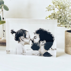 May include: A black and white photograph of a man and a child touching noses, encased in a clear acrylic block. The image is displayed on a white surface, with decorative plants in the background.