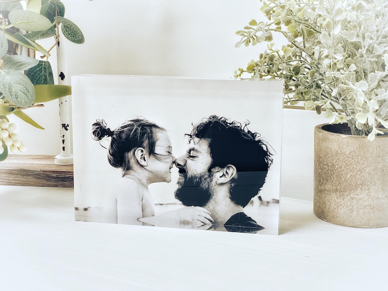 May include: A black and white photograph of a man and a child touching noses, encased in a clear acrylic block. The image is displayed on a white surface, with decorative plants in the background.