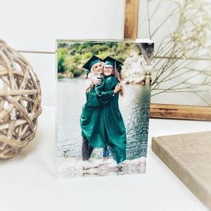 May include: A clear acrylic photo block featuring a graduation photo of two people in green gowns and black caps, embracing. The background shows a lake and trees. A decorative wooden ball and framed art are in the background.