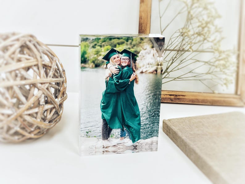 May include: A clear acrylic photo block featuring a graduation photo of two people in green gowns and black caps, embracing. The background shows a lake and trees. A decorative wooden ball and framed art are in the background.