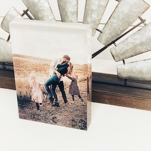 May include: A rectangular photo print featuring a family embracing in a field. The print is displayed on a wooden base, with a decorative metal windmill in the background. The photo captures a moment of affection and joy.