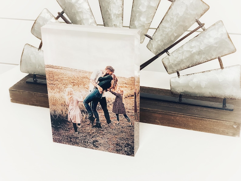 May include: A rectangular photo print featuring a family embracing in a field. The print is displayed on a wooden base, with a decorative metal windmill in the background. The photo captures a moment of affection and joy.