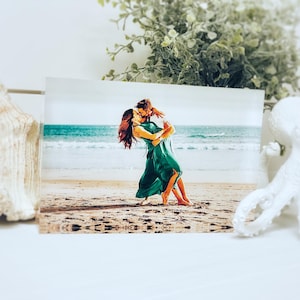 May include: A rectangular print featuring a couple embracing on a beach, with the ocean in the background. The couple is wearing green dresses. A white seashell and a white octopus figurine are in the foreground.