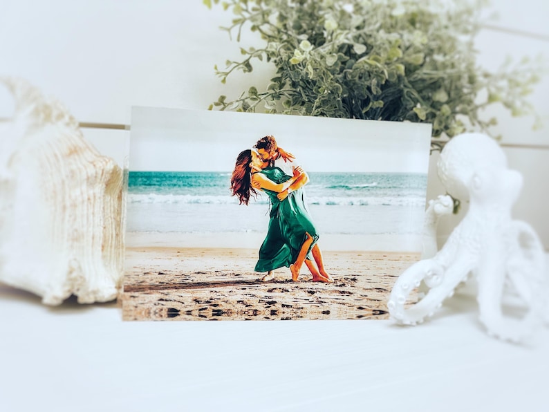 May include: A rectangular print featuring a couple embracing on a beach, with the ocean in the background. The couple is wearing green dresses. A white seashell and a white octopus figurine are in the foreground.