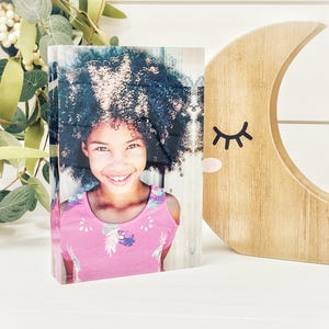 May include: A photo block featuring a smiling girl with curly hair wearing a pink top. A wooden crescent moon with a closed-eye design is to the right. Greenery with white berries is in the background.