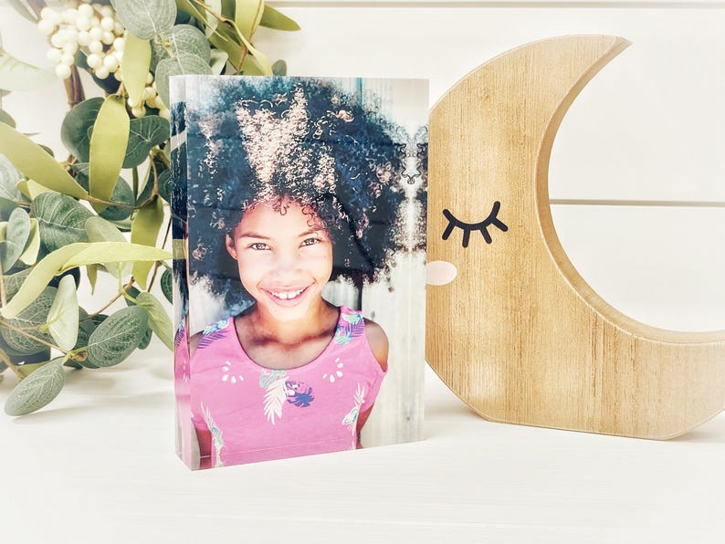 May include: A photo block featuring a smiling girl with curly hair wearing a pink top. A wooden crescent moon with a closed-eye design is to the right. Greenery with white berries is in the background.