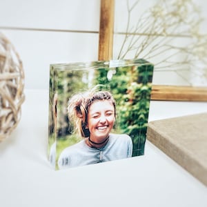 May include: A clear acrylic photo block featuring a smiling person in a gray shirt. The image is set against a blurred green background. A decorative sphere and a wooden frame are in the background.