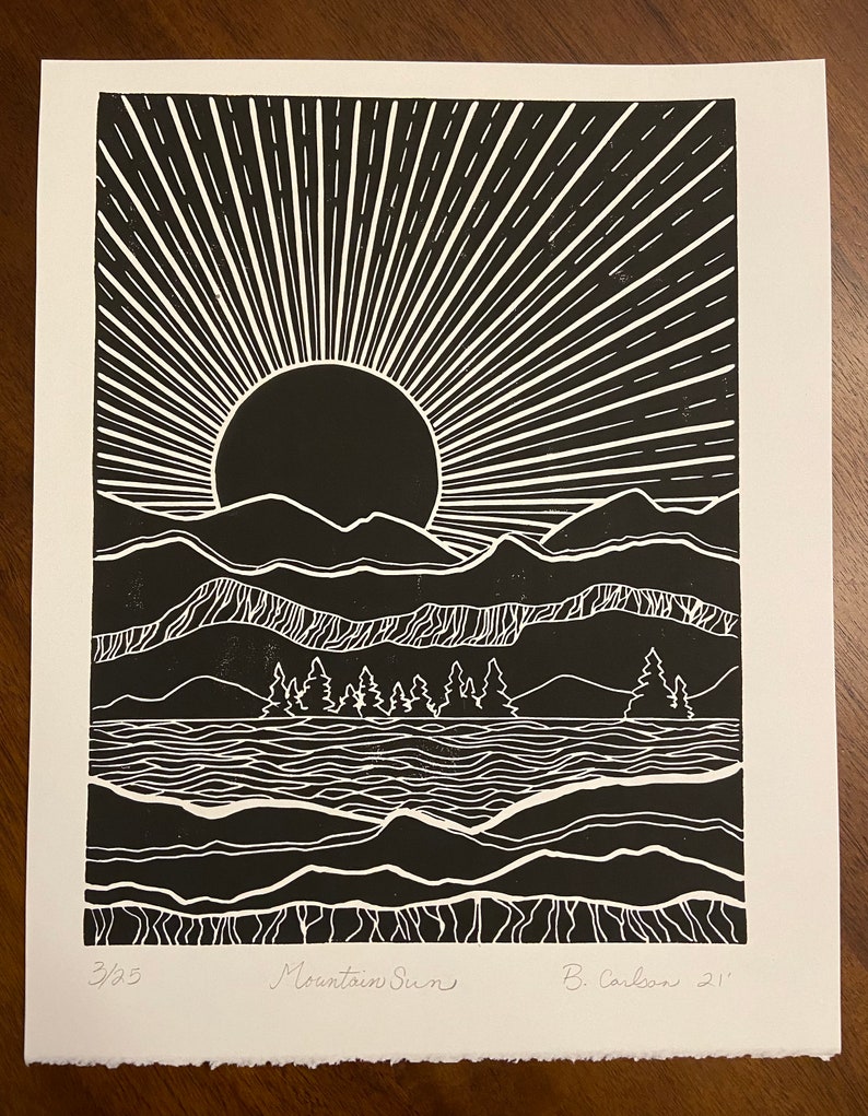 May include: Black and white linocut print of a mountain scene with a sun rising over a lake. The print is titled "Mountain Sun" and signed "B. Carbon 21'" in the bottom right corner. The print is numbered 3/25 in the bottom left corner.