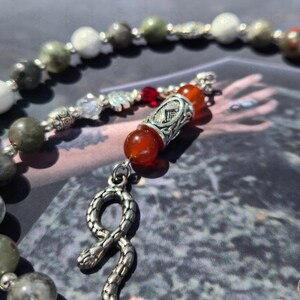 May include: A beaded necklace featuring a silver-toned snake pendant, interspersed with round white, grey, and green stone beads. The design includes a central silver-toned metal bead with an etched symbol, flanked by two translucent orange beads.