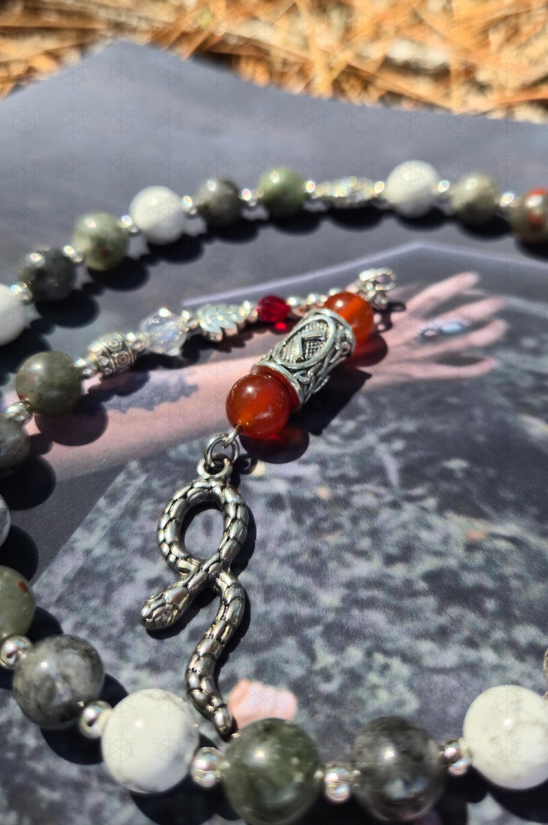 May include: A beaded necklace featuring a silver-toned snake pendant, interspersed with round white, grey, and green stone beads. The design includes a central silver-toned metal bead with an etched symbol, flanked by two translucent orange beads.