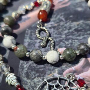 May include: A beaded necklace featuring white, grey, and green stone beads interspersed with small silver-toned metal spacers. The necklace includes a silver-toned snake charm, a red bead, and a silver-toned circular pendant with a web-like design.