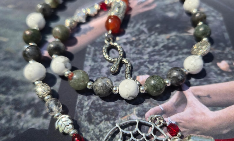 May include: A beaded necklace featuring white, grey, and green stone beads interspersed with small silver-toned metal spacers. The necklace includes a silver-toned snake charm, a red bead, and a silver-toned circular pendant with a web-like design.