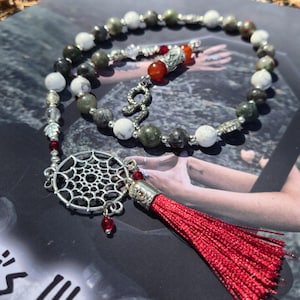 May include: A beaded necklace featuring alternating white and dark grey stone beads, silver-toned metal spacers, and a central silver-toned dreamcatcher charm. The piece includes a bright red tassel, small red accent beads, and a snake-shaped charm.