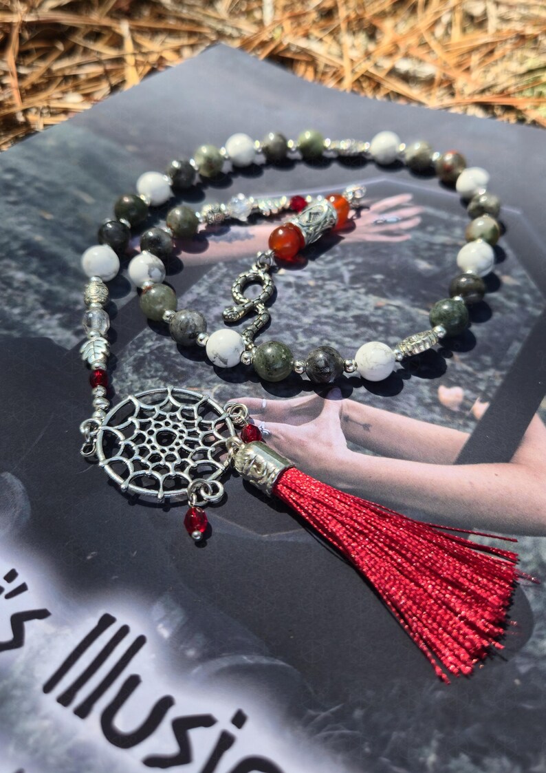 May include: A beaded necklace featuring alternating white and dark grey stone beads, silver-toned metal spacers, and a central silver-toned dreamcatcher charm. The piece includes a bright red tassel, small red accent beads, and a snake-shaped charm.