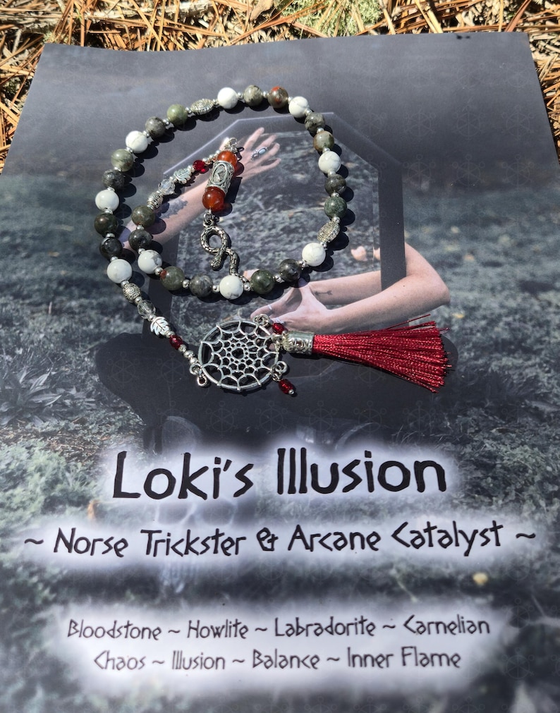 May include: A beaded necklace featuring green, white, and grey stone beads, silver-toned metal accents, a silver dreamcatcher charm, and a red tassel. The necklace rests on a card labeled Loki's Illusion, Norse Trickster & Arcane Catalyst.