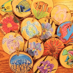 May include: A collection of hand-painted wooden ornaments featuring marine life and floral designs. The ornaments include images of fish, sea turtles, jellyfish, and flowers, each with a natural wood grain background and twine for hanging.