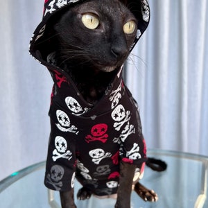 May include: A black hairless cat wearing a black hooded sweatshirt with a skull and crossbones pattern. The hoodie has red and white accents.