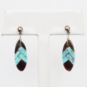 May include: A pair of silver-toned earrings with an oval, inlaid design. The earrings feature a combination of turquoise and dark brown stones in a geometric pattern. The earrings are displayed on a white stand.