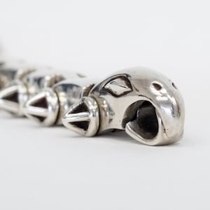 May include: Close-up of a silver-toned metal bracelet featuring a series of stylized animal heads and spiked accents. The bracelet has a polished finish and a detailed design, with a focus on the animal head's open mouth.