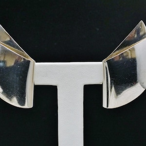 May include: A pair of silver-toned earrings with a geometric design. Each earring features a curved, fan-like shape with a folded, layered appearance. The earrings are displayed on a white stand against a black background.
