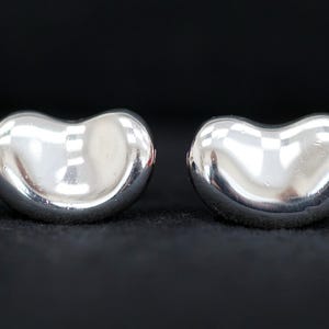 May include: A pair of polished silver bean-shaped stud earrings. The earrings have a smooth, reflective surface, and are set against a dark background. The earrings are a classic jewelry design.