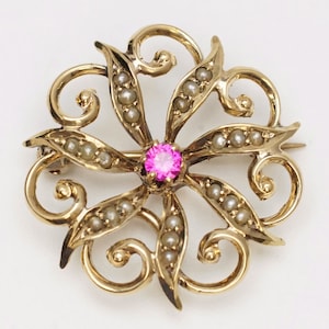 May include: A gold brooch with a pink gemstone in the center and a swirling design of pearls around it.