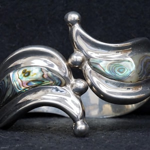 May include: A silver cuff bracelet with an abstract design. The bracelet features two curved sections, each inlaid with iridescent abalone shell. The design includes small, spherical accents and is set against a dark background.