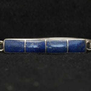 May include: A silver bracelet featuring four rectangular, deep blue lapis lazuli stones. The stones are set in a silver frame, creating a striking contrast. The bracelet has a simple, elegant design, perfect for everyday wear.