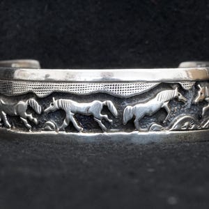 May include: Silver cuff bracelet featuring a detailed relief of running horses. The horses are depicted in a stylized manner, set against a backdrop of rolling hills and a textured sky. The bracelet has a polished finish and a classic, open design.