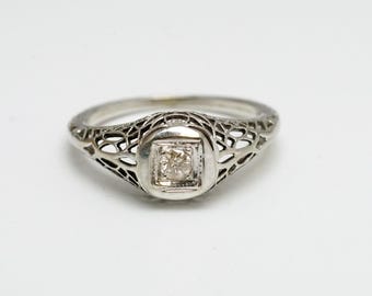 May include: A silver filigree ring with a single, clear, square-cut diamond in the center.