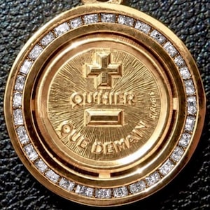 May include: A round gold pendant featuring a border of small clear stones. The centre displays a raised cross, a horizontal bar, and the French text "QU'HIER QUE DEMAIN" with the word "AUGIS" vertically inscribed on the right side of the disc.
