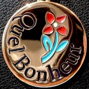 Puede incluir: Un colgante dorado con la frase "Que le Bonheur" en letras negras. El colgante presenta una flor estilizada con pétalos rojos, un centro blanco y hojas azules. El fondo es negro.