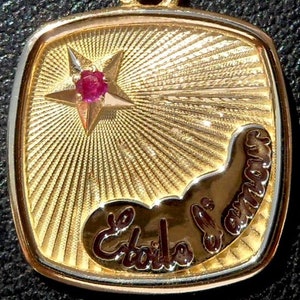 Könnte beinhalten: Ein goldfarbener Anhänger mit quadratischer Form und Sternenmuster. Ein fünfzackiger Stern mit einem roten Edelstein befindet sich über dem Text "Étoile d'amour" in Schreibschrift. Der Anhänger hat einen goldenen Rand.